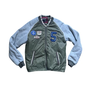 Grön bomberjacka - Snygg grön bomberjacka från Jack & Jones med grå ärmar och coola patches på bröstet. Jackan har dragkedja framtill, ribbade muddar och kontrastfärgade detaljer. Perfekt för dig som gillar sportig och avslappnad stil.