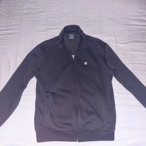 Champion ziptröja  - Kom med pris. Svart ziptröja från Champion med klassisk logga på bröstet. Tröjan har hög krage, hel dragkedja och två fickor framtill. Tillverkad i mjuk bomullsblandning, perfekt för lager-på-lager och chill dagar.