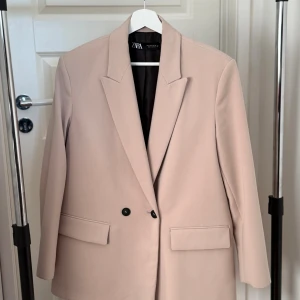 Beige kavaj från Zara, storlek S - Stilren beige kavaj från Zara med klassisk krage och två knappar framtill. Kavajen har raka fickor med lock och är helfodrad med svart innerfoder. Perfekt för en clean och modern look.