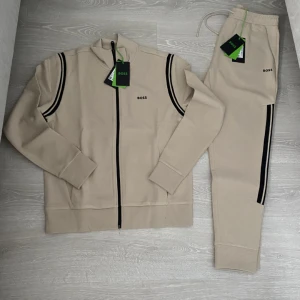 Boss Tracksuit  - (Värde på 3600) Snygg beige ziptröja från BOSS med svarta ränder längs axlarna och dragkedja framtill. Tröjan har hög krage och ribbade muddar. Perfekt för en clean och sportig look. Matchande byxor finns också.