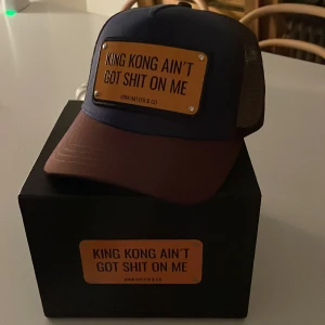 John Hatter & Co keps med statement - Snygg keps från John Hatter & Co med mörkblå front, brunt skärm och mesh i brunt. Framtill sitter en metallskylt med texten 'KING KONG AIN'T GOT SHIT ON ME'. Justerbar baktill och levereras med originalpåse.Helt ny,med prislapp