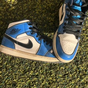 Nike Air Jordan 1 blå/vita sneakers - Säljer ett par Nike Air Jordan 1 sneakers i blått, vitt och svart. Klassisk high-top modell med svart swoosh, svarta skosnören och perforerad tå. Skorna är i skinn och har en ikonisk Jordan-logga på sidan. Perfekta för dig som vill sticka ut med en tidlös streetstyle.