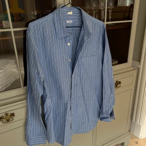 Blå randig skjorta i bomull - Säljer en blå skjorta från Uniqlo med vita ränder. Skjortan har klassisk krage, lång ärm och knäppning framtill. Tillverkad i mjuk bomull och passar perfekt till jeans eller chinos. Enkel bröstficka och relaxed fit.