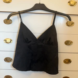 Svart blommig linnetopp med smala band - Snygg svart linnetopp med subtilt blommönster och smala axelband. Toppen har v-ringad hals och är croppad, vilket ger en trendig look. Materialet är glansigt och känns lyxigt, perfekt för att lyfta vilken outfit som helst.