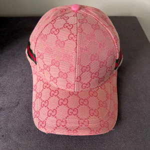 Rosa keps från Gucci med GG-mönster - Säljer en rosa keps från Gucci med ikoniskt GG-monogram över hela kepsen. Kepsen har justerbart spänne bak i rosa läder och klassiska gröna och röda ränder på sidan. Tillverkad i bomull och detaljer i läder. Perfekt för dig som vill sticka ut med en lyxig accessoar.