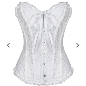 Vit korsett med rosett och spets - Supersnygg vit korsett med dekorativ rosett framtill och fina spetsdetaljer längs nederkanten. Korsetten har ett subtilt mönster och knäppning framtill, perfekt till halloween! Säljer även en vit gloria och änglavingar!