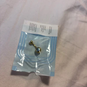 Guld navelpiercing med glittrig sten helt oanvänd!  - Helt oanvänd snygg navelpiercing från India Bodypiercing & Jewellery i kirurgiskt stål med guldtonad finish. Tappar inte färg! Hade råkat beställa 2 st därför jag säljer vidare och har använd den andra vilket är superbra kvalité. Piercingen har två klara, glittriga stenar som fångar ljuset och ger extra bling. Perfekt för dig som vill ha något som sticker ut och känns lyxigt. 10 mm