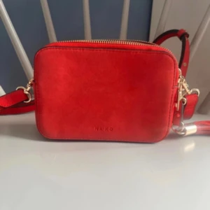 Röd väska - Snygg röd axelväska från NA-KD i mjuk mocka med två stora fack och guldfärgade dragkedjor. Justerbar axelrem med coola nitar och dekorativ tofs. Perfekt storlek för mobilen och det viktigaste. Enkel, stilren design som sticker ut.