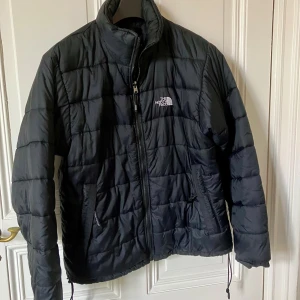 Svart pufferjacka från The North Face - Svart pufferjacka från The North Face med quiltad design och hög krage. Jackan har dragkedja framtill, två sidofickor med dragkedja och justerbar dragsko nedtill. Yttermaterial i nylon och vaddering i polyester. Perfekt för kalla dagar.