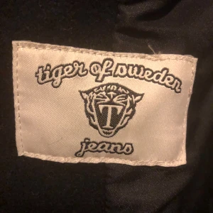 Svart skepparkavaj från Tiger of Sweden - Stilren svart skepparkavaj från Tiger of Sweden med dubbla knapprader och snygga detaljer i skinn vid fickorna. Jackan har bred krage och klassisk marin känsla, perfekt för dig som vill ha en tidlös look med edge.