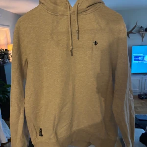 Beige hoodie från Morris Stockholm - Snygg beige hoodie från Morris Stockholm med huva och snörning. Tröjan har en liten broderad logga på bröstet och är tillverkad i mjuk bomull. Perfekt för chill dagar och enkel att matcha med jeans eller joggers. Finns inga slitningar i hoodien, anledning till att den ser skrynklig ut är för den är ny tvättad!