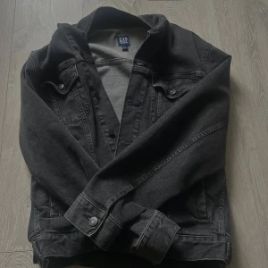 Svart jeansjacka från GAP, storlek S - Snygg svart jeansjacka från GAP i klassisk modell med knappar framtill och bröstfickor med lock. Jackan har en rak passform och är tillverkad i slitstarkt denimtyg. Perfekt för dig som gillar en stilren och tidlös look. Priset går alltid att diskutera!