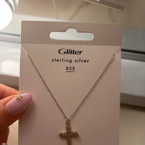 Silverhalsband med kors  - Snyggt halsband i äkta sterling silver 925 från Glitter. Kedjan är tunn och smidig, med ett hängsmycke i form av ett kors dekorerat med små glittrande stenar. Perfekt accessoar för att lyfta din outfit med en stilren touch.