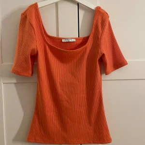 Ribbad orange  topp från Pieces - Säljer en ribbad orange topp från Pieces i storlek XS. Toppen har en fyrkantig ringning och korta ärmar. Materialet är stretchigt och ger en snygg passform. Perfekt att styla med jeans eller kjol för en färgstark look. Man kan även ha den som en off shoulder topp🧡
