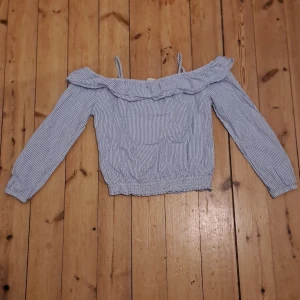 Randig offshoulder blus från H&M - Säljer en söt blus från H&M i storlek 140. Blusen är vit med tunna blå ränder, har offshoulder-modell med volang upptill och smala axelband. Långärmad och med resår i midjan och vid ärmsluten. Perfekt för vår och sommar!