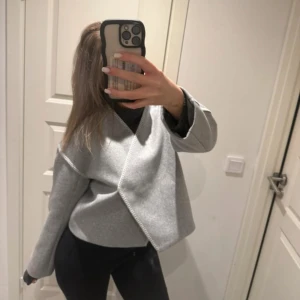 Ljusgrå omlott kappa i ullmix - Snygg ljusgrå kappa med omlott-design och markerade sömmar. Jackan har en boxig, loose passform och är tillverkad i ullmix som ger en mjuk och stilren känsla. Perfekt för dig som gillar minimalistisk och clean stil.