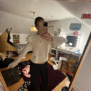 Vit stickad blus från Holly & Whyte - Supersöt vit stickad blus från Holly & Whyte med volangdetalj över axlarna och diskreta lodräta ränder. Blusen har långa ärmar och en lätt croppad passform, perfekt att styla med höga jeans eller kjol. Passar S oxåå 