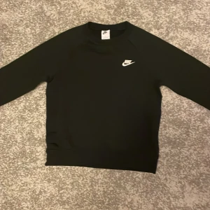 Svart Nike sweatshirt med logga - Svart sweatshirt från Nike med klassisk vit broderad logga på bröstet. Tröjan har rund halsringning och långa ärmar. Perfekt basic-plagg med stilren look och mjukt material.
