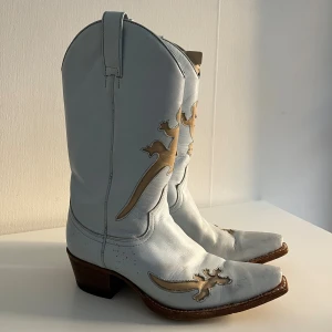 Ljusblå cowboy boots i skinn, stl 37 - Unika ljusblå cowboy boots från Sendra i äkta skinn med beige dekorativa detaljer i form av ödlor och spetsig tå. Snygg kontrastsöm och klassisk hög klack. Skorna är väl använda och har därav slitage i form av mindre repor och fläckar - men kan rengöras något innan de skickas! 