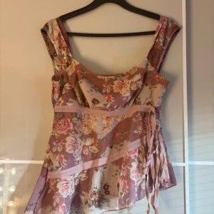 Blommig sommar blus i rosa toner - Supersöt kort blommig top  i rosa och beige toner med omlott och knytband i sidan.  har breda axelband och ett flowy fall, perfekt för varma dagar. Materialet känns lätt och luftigt, och mönstret är romantiskt med stora blommor.