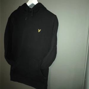Svart hoodie från Lyle & Scott - Svart hoodie från Lyle & Scott med klassisk gul logga på bröstet. Tröjan har huva med snörning, känguruficka och långa ärmar. Tillverkad i mjuk bomull, perfekt för chill dagar eller när du vill ha en clean look.