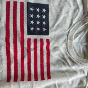 Vit stickad tröja med USA-flagga Gant - Vit stickad tröja från Gant med stor amerikansk flagga på bröstet i rött, blått och vitt. Rund halsringning och klassisk passform. Perfekt för dig som vill sticka ut med en preppy look.