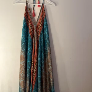 Färgglad klänning  - Lång, ärmlös klänning med v-ringning och knytband i nacken och ryggen. Klänningen har ett vackert mönster i blått och brunt med detaljer i orange. Den har även dekorativa tofsar. Klänningen går att knyta själv runt nacke och rygg så storleken är free size, man bestämmer lite själv, så allt från S-XL beroende på hur man knyter den. Nypris 42 euro, köpt utomlands och aldrig använd 