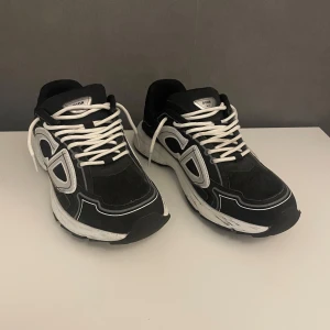 Svarta Dior sneakers med vita detaljer - Säljer ett par svarta Dior sneakers med vita och silverfärgade detaljer. Skorna har tydliga Dior-loggor på sidan och bak, samt vita snören och en chunky vit sula. Ovandelen är i mesh och syntetmaterial för en sportig vibe. Perfekta för dig som vill sticka ut med lyxig streetstyle. Säljs billigt pga ingen kartong eller kvitto! 