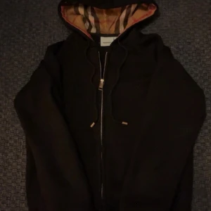 Svart Burberry hoodie med dragkedja - Svart hoodie från Burberry med klassiskt rutigt foder i huvan och broderad logga på bröstet. Tröjan har dragkedja framtill, snörning i huvan och guldfärgade detaljer på snörena. Perfekt för dig som vill ha en stilren och lyxig look.pris kan diskuteras!
