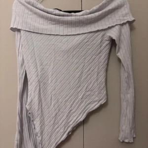 Vit ribbad offshoulder topp från AWARE - Säljer en vit ribbad offshoulder topp från AWARE med lång ärm och asymmetrisk nederkant. Toppen har en bred krage som vilar snyggt över axlarna och är tillverkad i ett mjukt, stretchigt material. Perfekt för dig som vill sticka ut med en unik siluett.