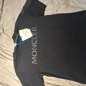 Svart Moncler t-shirt med logga - Svart t-shirt från Moncler med stor logga tryckt vertikalt på framsidan. Klassisk rund halsringning och korta ärmar. Ena ärmen har en ficka med knapp och Moncler-märke. Tillverkad i mjuk bomull för en skön känsla. HELT NY OCH ÄKTA
