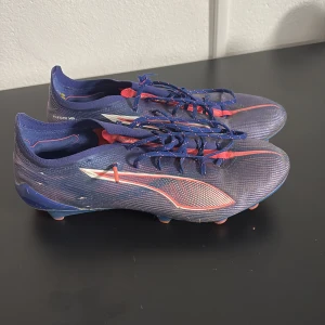 Blåa Puma fotbollsskor med orange detaljer - Säljer ett par snygga blåa Puma fotbollsskor med orange och röda detaljer. Skorna har snörning och en lätt, syntetisk ovandel samt dobbar på sulan för bra grepp på planen. Perfekta för dig som vill sticka ut på fotbollsplanen!