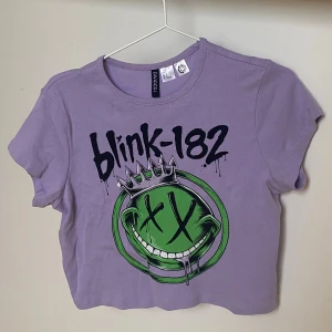 Lila Blink-182 croppad t-shirt H&M - Säljer en lila croppad t-shirt från H&M Divided med Blink-182 tryck . T-shirten har korta ärmar och ett stort grönt smileytryck med krona. Går att fråga om prissänkning. Tvättas innan den skickas