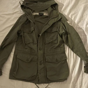 Ralph lauren Fieldjacka  - Snygg grön parkajacka med huva och flera praktiska fickor. Jackan har en cool broderad detalj på ärmen och stängs med både dragkedja och knappar. Perfekt för höst och vår.