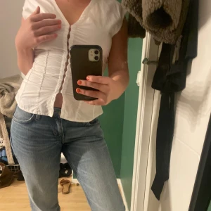 Vit blus med knappar och rynk - Säljer en vit blus med v-ringning och snygga rynkdetaljer längs sidorna. Blusen har korta ärmar och knappar framtill som ger en trendig look. Perfekt att styla med jeans eller kjol för en fräsch outfit. Köptes för 400 från början❤️