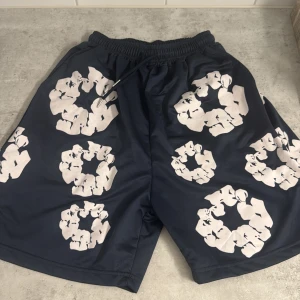 Mörkblå shorts från Denim Tears - Säljer ett par mörkblå shorts från Denim Tears med unika vita blommönster över hela plagget. Shortsen har elastisk midja med snörning och en bakficka. Perfekta för dig som vill sticka ut med en streetwear vibe.