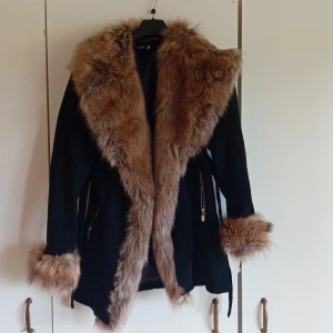 Svart pälsjacka med stora bruna detaljer - Snygg svart pälsjacka med stora, fluffiga bruna fuskpälsdetaljer vid krage, ärmslut och längs öppningen. Jackan har dragkedjor vid fickorna och ett bälte i midjan för extra stil. Perfekt för dig som vill sticka ut med en statementjacka i vinter.