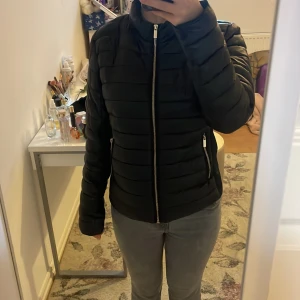 Svart pufferjacka från MNG, storlek M - Snygg svart pufferjacka från MNG i storlek M. Jackan har quiltad design, raka dragkedjor i guld på fickor och framtill. Materialet är glansigt och känns lätt, perfekt för kyliga dagar. Stilren och enkel att matcha med allt.
