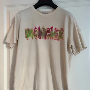 Beige Moncler t-shirt med färgglatt tryck - Snygg beige t-shirt från Moncler med stort färgglatt logotryck i grönt och rosa på bröstet.  Storlek 164/14y 