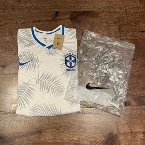 Vit Brasilien fotbollströja Nike - Snygg vit Brasilien fotbollströja från Nike med grå och blå palmbladsmönster. Tröjan har blå detaljer vid krage och ärmslut, samt broderat Brasilien-märke på bröstet. Tillverkad i lätt och ventilerande material, perfekt för match eller träning.