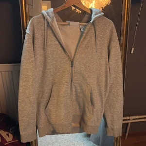 Grå hoodie med dragkedja från Weekday - Mysig grå hoodie från Weekday i storlek S. Tröjan har dragkedja framtill, huva med snörning och stora fickor. Perfekt för chill dagar och enkel att matcha med jeans eller mjukisbyxor.