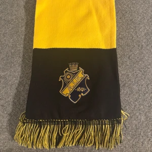 AIK halsduk svart och gul med tofsar💛🖤 - Grym halsduk i svart och gul med AIK:s broderade klubbmärke och årtalet 1891. Klassisk blockrandig design och snygga tofsar i ändarna. Perfekt accessoar för att visa ditt AIK-stöd på matchen eller i vardagen.