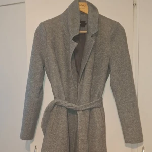 Grå kappa med bälte - Stilren grå kappa med bälte i midjan och hög krage. Kappan har lång ärm och en klassisk rak passform som ger en snygg siluett. Perfekt för dig som vill ha en enkel men trendig look under kyliga dagar.