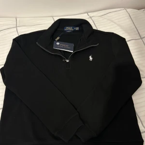 Svart half zip tröja Polo Ralph Lauren - Svart långärmad tröja från Polo Ralph Lauren med klassisk vit broderad logga på bröstet. Tröjan har en snygg half zip-dragkedja och hög krage. Perfekt för dig som gillar stilren och enkel design. Materialet är mjukt och skönt bomull.