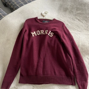 Vinröd stickad tröja från Morris - Vinröd stickad tröja från Morris med broderad logga i vitt framtill och Union Jack-detalj på ärmen. Klassisk rund halsringning och ribbade muddar vid ärmslut och nederkant. Perfekt för dig som gillar stilren och preppy look.