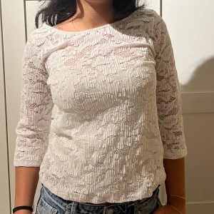 Vit spetsblus med trekvartsärm - Säljer en söt vit blus i spets med trekvartsärmar och rund halsringning. Blusen har ett diskret mönster och är lätt transparent, perfekt att matcha med jeans eller kjol. Den har en feminin och fräsch look som passar till många stilar.