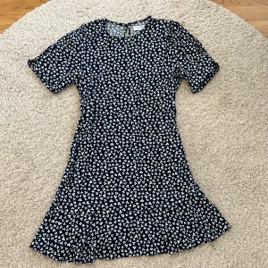 Blommig kortklänning från Holly & Whyte - Klänning från Holly & Whyte med mörkblå botten och vita blommor. Klänningen har rund hals, korta ärmar och en liten knapp i nacken. Materialet är mjukt och lätt, perfekt för varma dagar☀️
