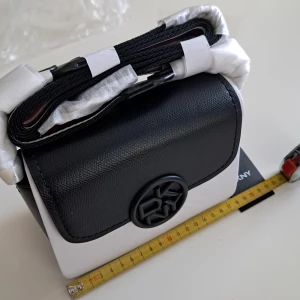 Svartvit axelväska från DKNY - Snygg svart och vit axelväska från DKNY med rund logotyp framtill. Väskan har en bred, randig axelrem i svart, vitt och vinrött samt stilren, boxig form. Läder, perfekt för dig som vill ha en trendig accessoar.