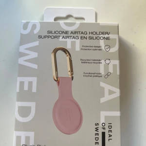 Silicone Airtag Holder Blush Pink - Elegant silikonhållare för Apple Airtag i blush pink med guldfärgad karbinhake. Skyddar din Airtag och är tillverkad av återvunnet material. Praktisk krok för enkel fastsättning på nycklar, väskor eller andra tillhörigheter. Produkten är ny i originalförpackning utan synligt slitage.