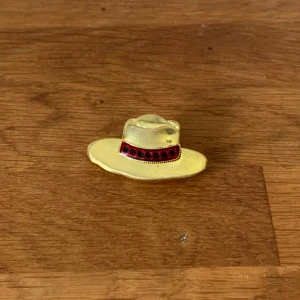 Cwboyhatt brosch med röda detaljer - Cool brosch i form av en cowboyhatt i guldfärgad metall med ett rött band och svarta prickar runt hatten. Perfekt accessoar för att ge din outfit en unik och lekfull touch.